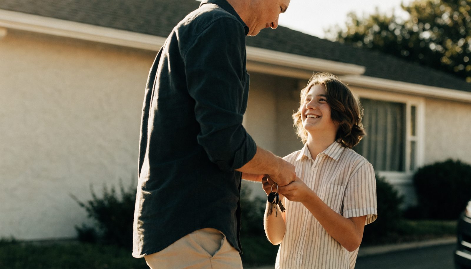 A parent handing car keys to a smiling teenage new driver in front of a California home